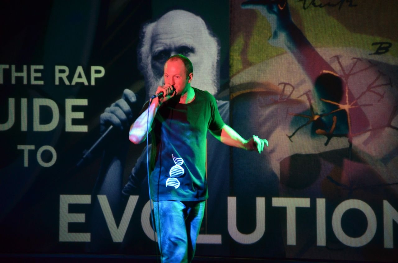 Baba Brinkman performing on stage in front of The Rap Guide to Evolution screen
