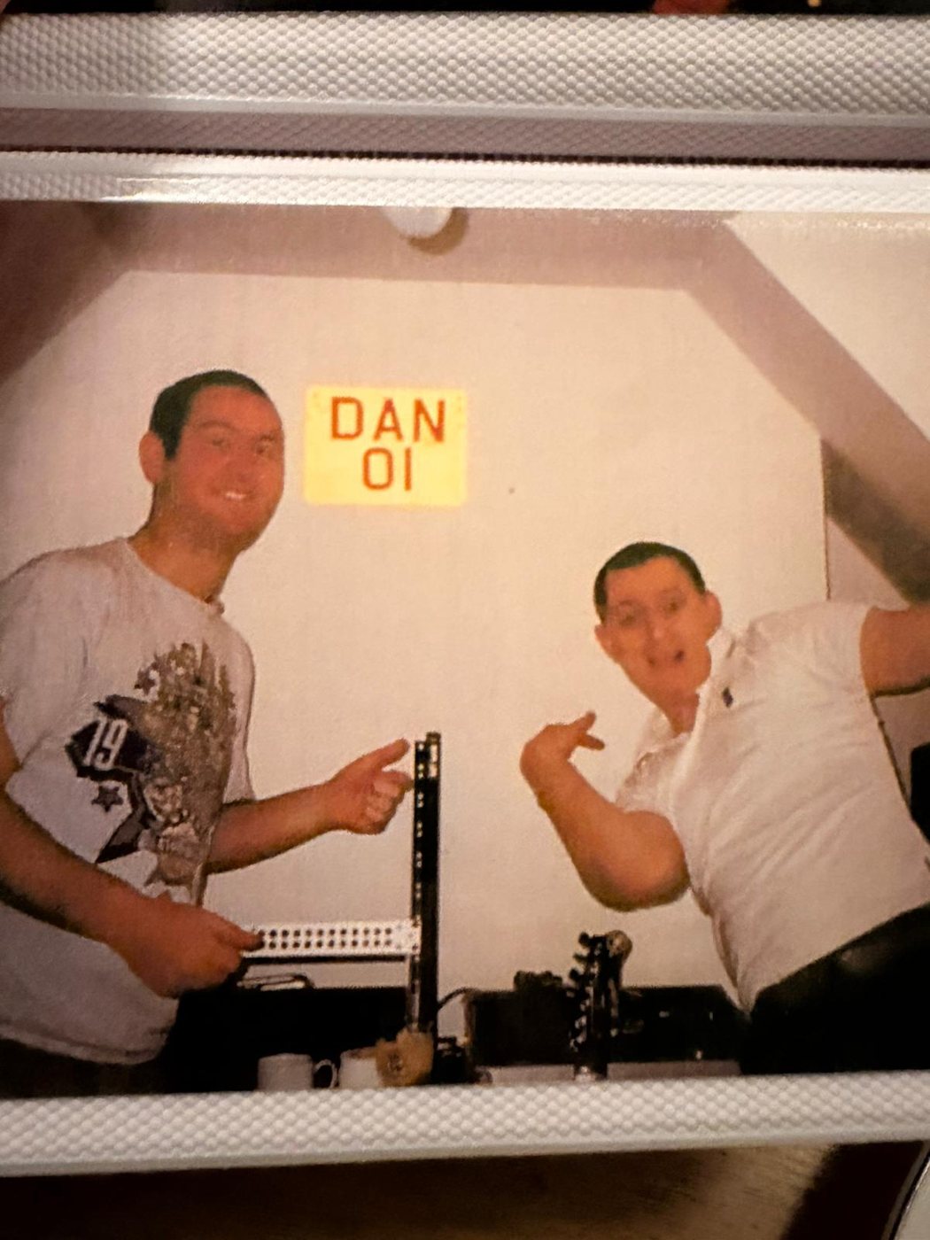Two people grinning in a studio under a DAN 01 sign, patch bay and guitar neck in front