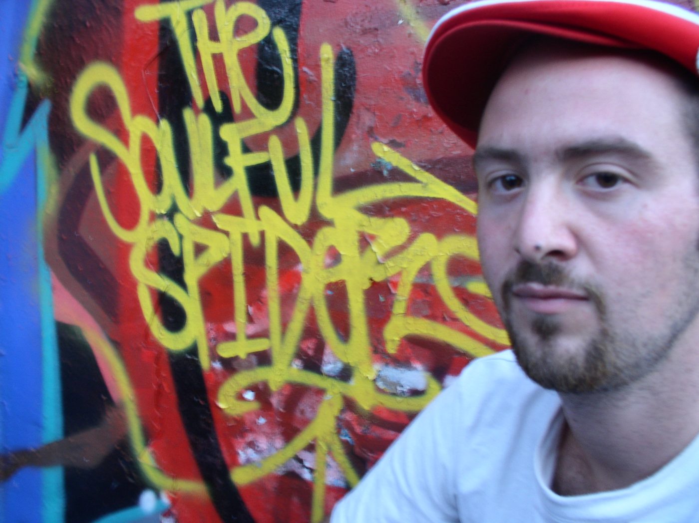 Close-up portrait in a red cap beside a yellow tag reading The Soulful Spider