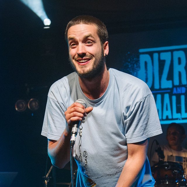 Dizraeli live on stage, smiling and leaning in with the mic, in front of a Dizraeli and the Small Gods screen
